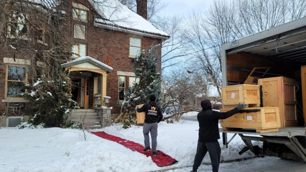 crown movers unloading a moving truck in and carrying boxes in over a red carpet laid across a snowy montreal front yard in the winter time.